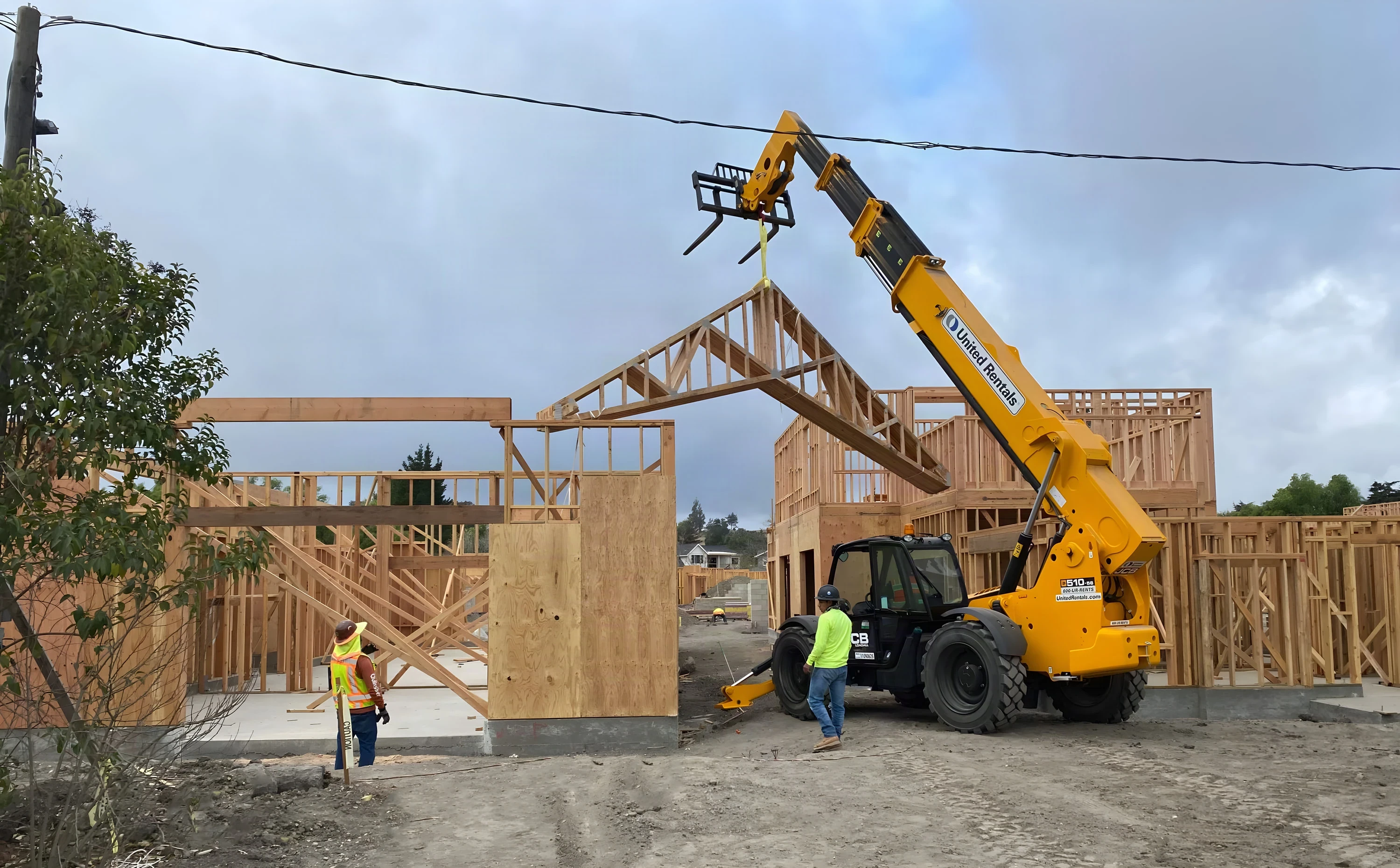 Crew and a telehandler setting wood roof trusses on a framed building under a blue sky