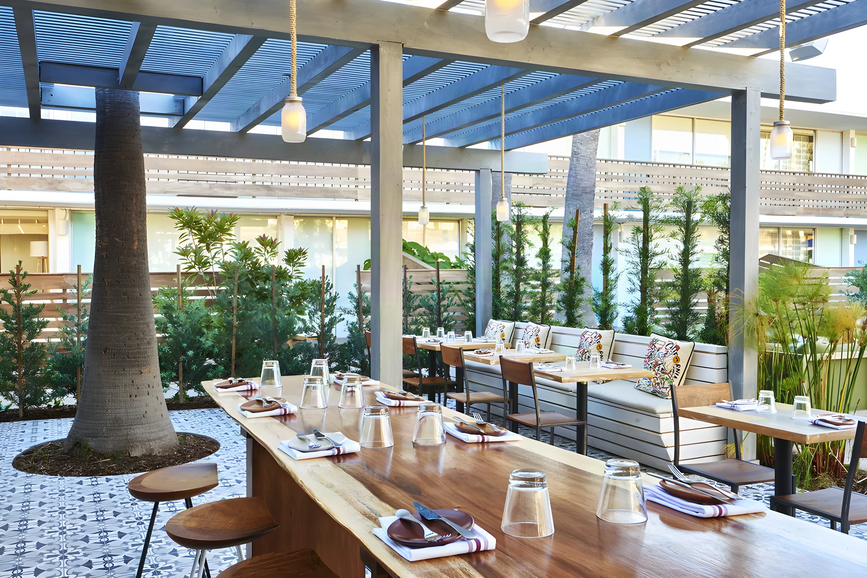 The Outpost patio with long timber tables under a blue slatted pergola and a tree