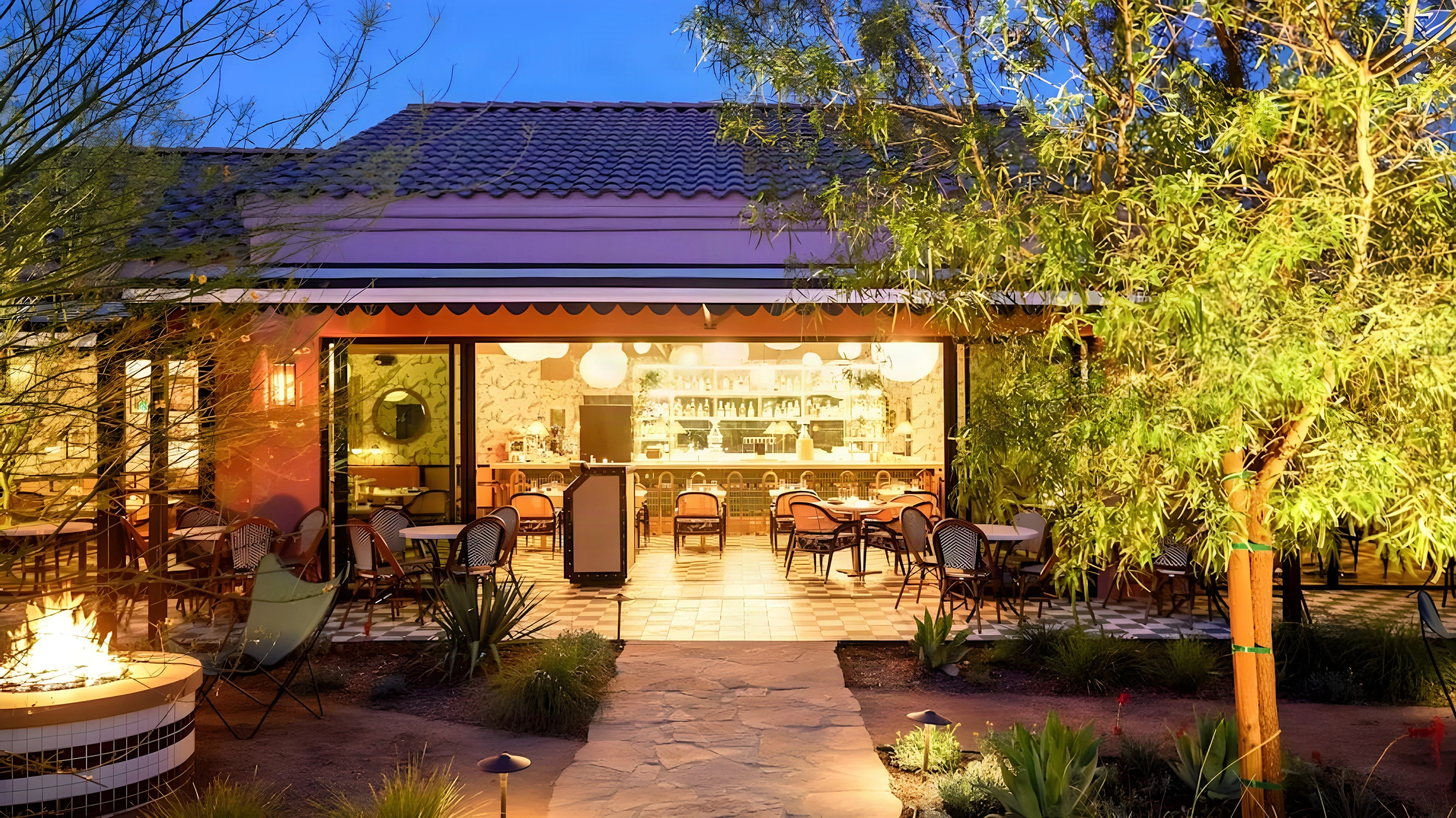 The Pink Cabana patio at dusk with a fire pit, desert trees and the lit dining room behind