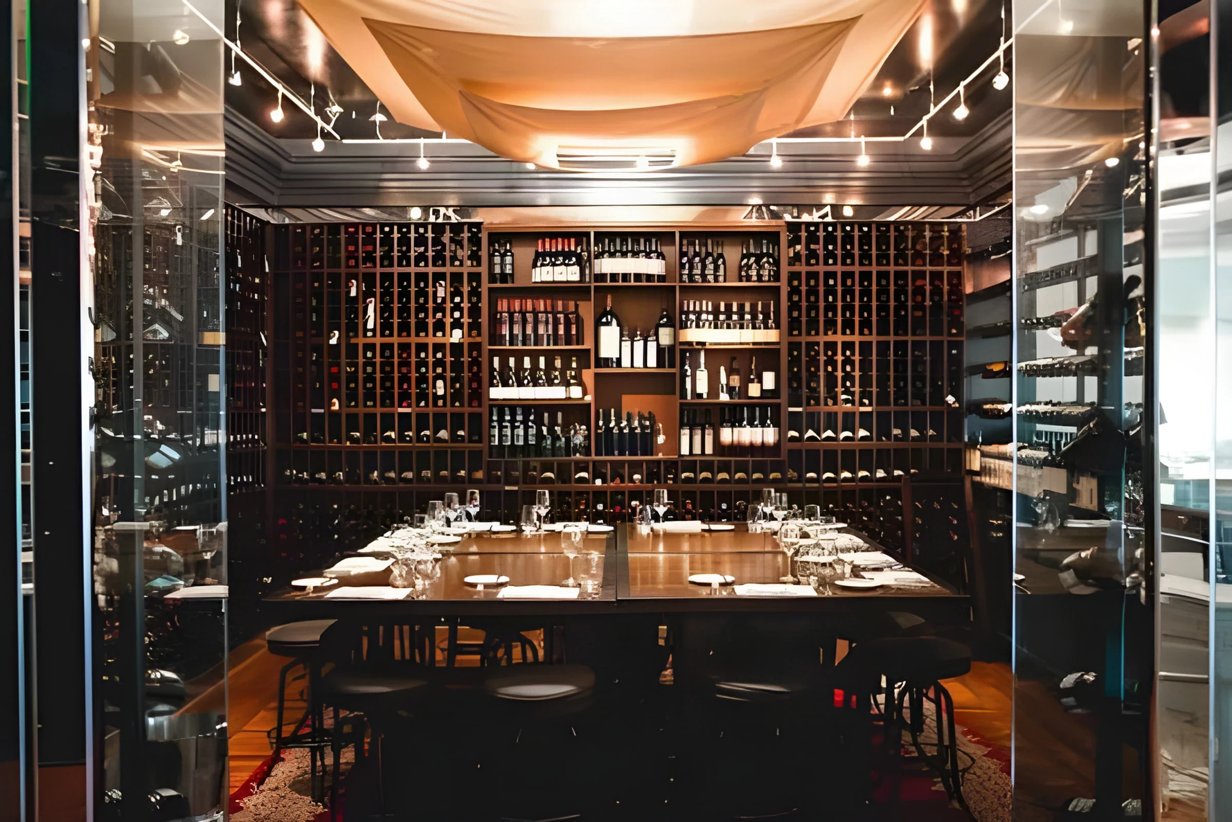 Table laid for dinner under track lighting, seen past the glass wall of the Royce Wine Room